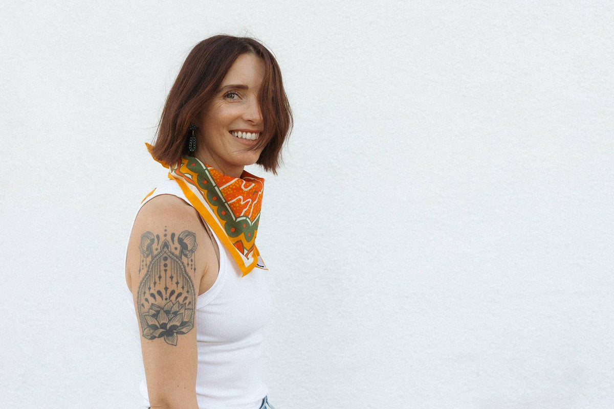 Smiling model wearing the Bandits Sunset bandana by Studio MUTI tied as a neckerchief, showcasing the bold orange and olive landscape-inspired artwork.
