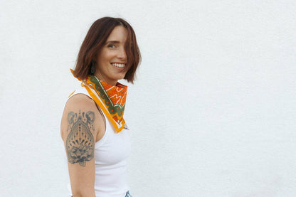 Smiling model wearing the Bandits Sunset bandana by Studio MUTI tied as a neckerchief, showcasing the bold orange and olive landscape-inspired artwork.