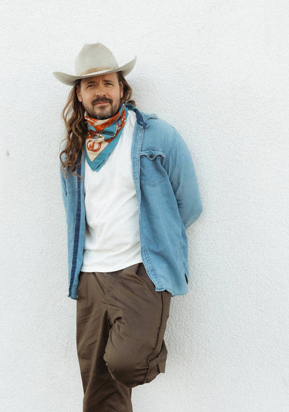 Model wearing the Bandits Wild West bandana by Van Berkemeyer tied as a neckerchief with a cowboy hat, denim overshirt, white T-shirt, and brown pants.