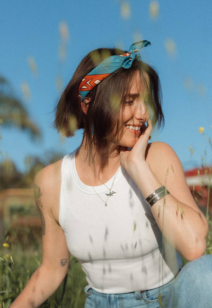Model wearing the Bandits Wild West bandana by Van Berkemeyer as a headband while sitting outdoors in a sunny field.