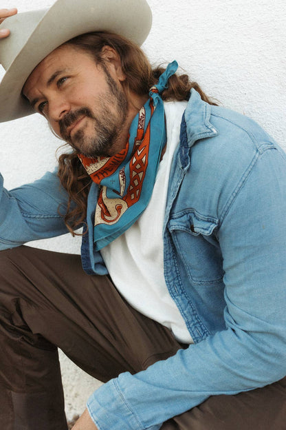 Model wearing the Bandits Wild West bandana by Van Berkemeyer tied as a neck scarf with a cowboy hat and denim overshirt.