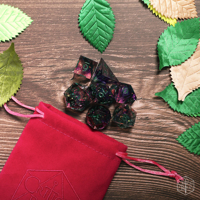 Critical Role Champion Dice Set inspired by Caduceus Clay displayed on a wood surface surrounded by green and cream leaves. The seven sharp-edged resin dice feature swirling pink, purple, and black tones with iridescent shimmer and green botanical inclusions, resting beside a dark pink Critical Role velveteen dice bag. The nature-inspired styling reflects Caduceus's connection to the Blooming Grove and the natural world.