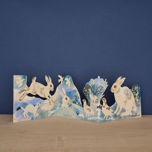 Mark Hearld Winter Wildlife accordion greeting card opened to display illustrated hares, ptarmigan, and a stoat in a snowy winter landscape.
