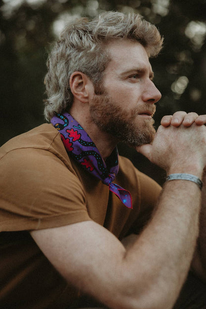 Model wearing the Pine Bones Electric Desert Parade bandana tied in a classic neckerchief style with the colorful desert wildlife artwork visible.