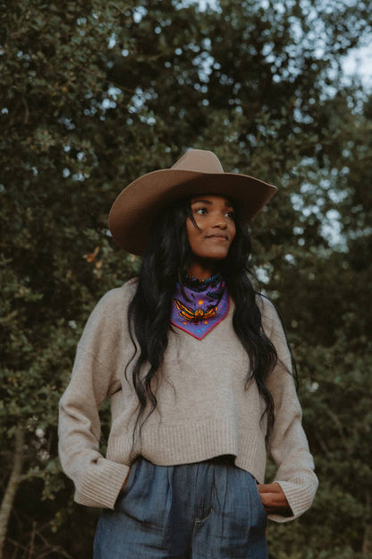 Model wearing the Pine Bones Electric Desert Parade bandana tied as a neck scarf with a tan hat, cream sweater, and denim trousers in an outdoor setting.