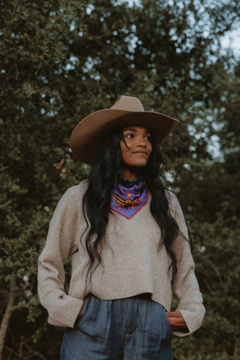 Model wearing the Pine Bones Electric Desert Parade bandana tied as a neck scarf with a tan hat, cream sweater, and denim trousers in an outdoor setting.