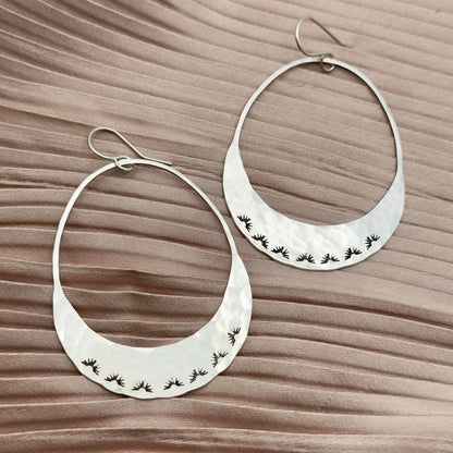 Fluff Hardware Lumos Earrings are handcrafted statement hoops with an organic shape and a one-of-a-kind stamped, textured finish. Designed to feel subtle yet striking, these small-batch earrings dangle from hypoallergenic sterling silver ear wires and add luminous movement to any outfit. A beautiful gift and an easy everyday accessory for anyone who loves modern, nature-inspired jewelry with artisan detail.