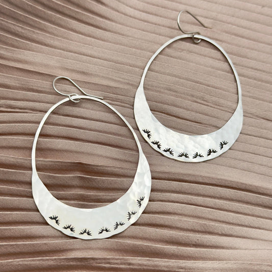 Fluff Hardware Lumos Earrings are handcrafted statement hoops with an organic shape and a one-of-a-kind stamped, textured finish. Designed to feel subtle yet striking, these small-batch earrings dangle from hypoallergenic sterling silver ear wires and add luminous movement to any outfit. A beautiful gift and an easy everyday accessory for anyone who loves modern, nature-inspired jewelry with artisan detail.