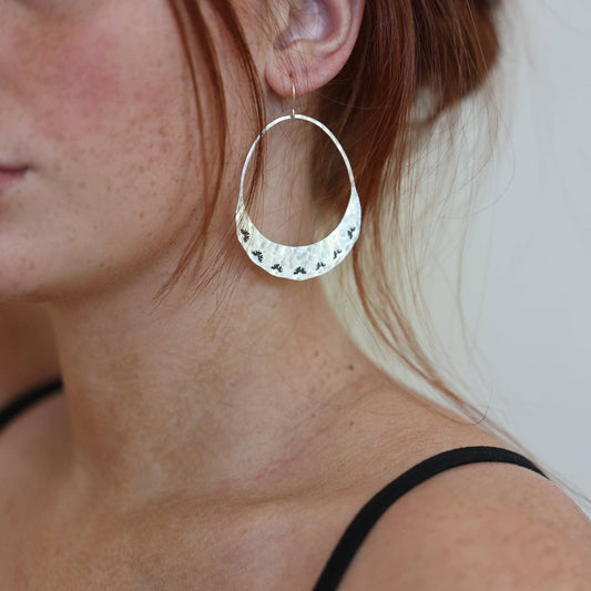 Fluff Hardware Lumos Earrings are handcrafted statement hoops with an organic shape and a one-of-a-kind stamped, textured finish. Designed to feel subtle yet striking, these small-batch earrings dangle from hypoallergenic sterling silver ear wires and add luminous movement to any outfit. A beautiful gift and an easy everyday accessory for anyone who loves modern, nature-inspired jewelry with artisan detail.