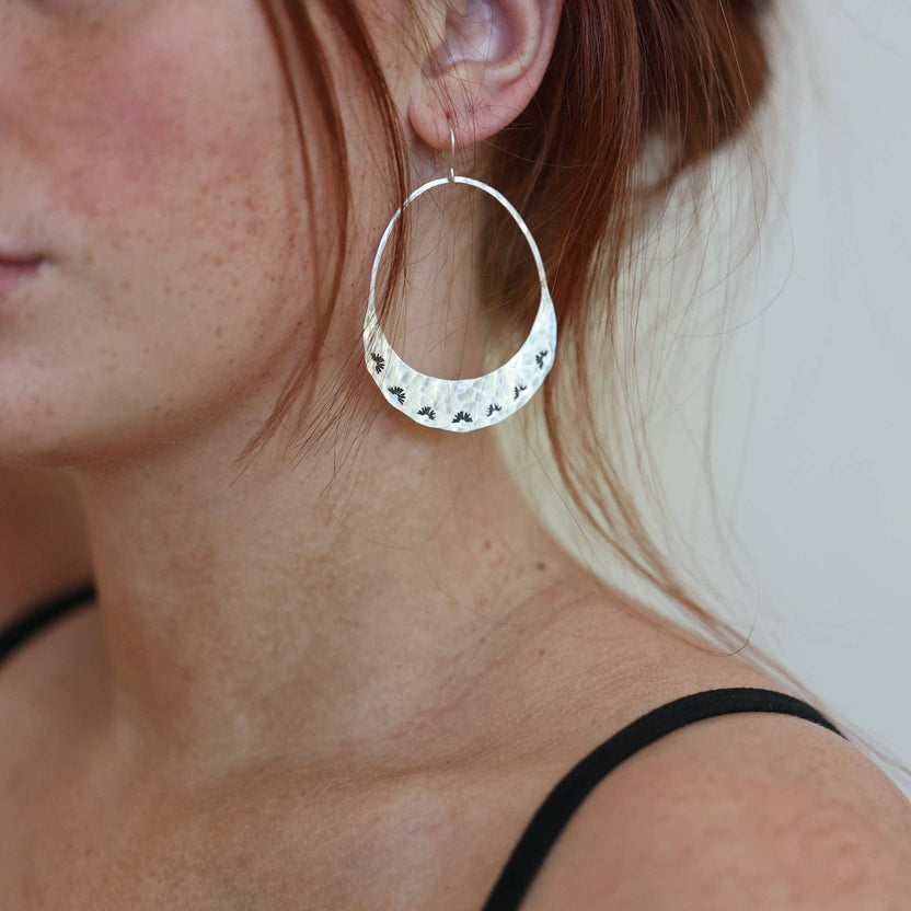 Fluff Hardware Lumos Earrings are handcrafted statement hoops with an organic shape and a one-of-a-kind stamped, textured finish. Designed to feel subtle yet striking, these small-batch earrings dangle from hypoallergenic sterling silver ear wires and add luminous movement to any outfit. A beautiful gift and an easy everyday accessory for anyone who loves modern, nature-inspired jewelry with artisan detail.