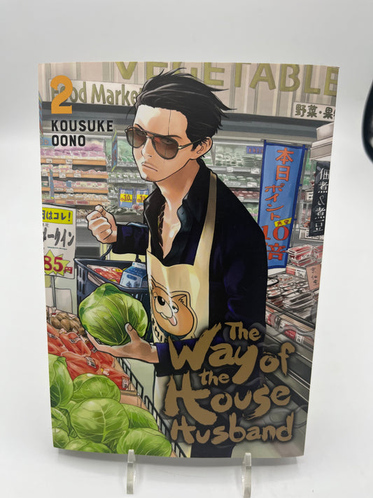 Cover of The Way of the Househusband Volume 2 by Kousuke Oono, featuring Tatsu wearing a cat apron and sunglasses while shopping for vegetables in a grocery store.