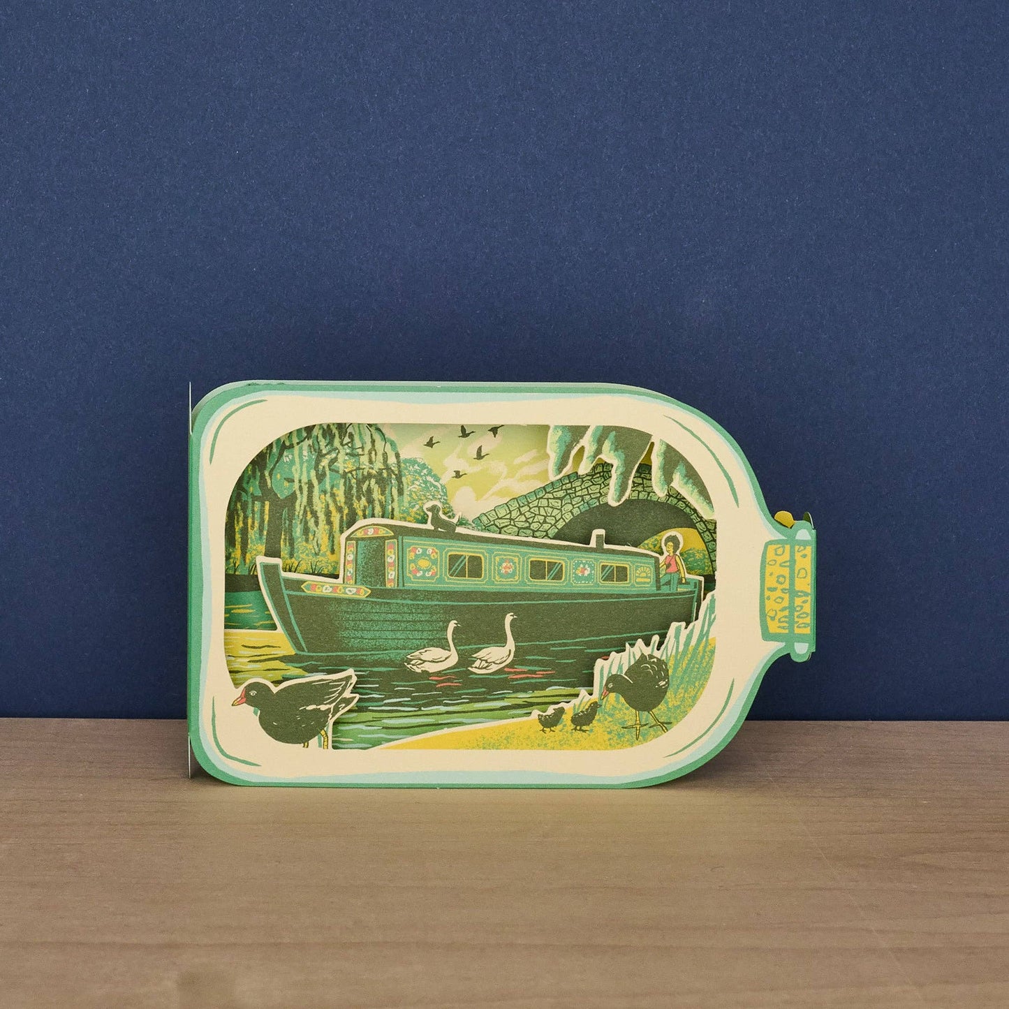 Front of Tom Jay Canal Boat in a Bottle die-cut greeting card shaped like a glass bottle featuring a colorful narrowboat, swans, and canal wildlife.