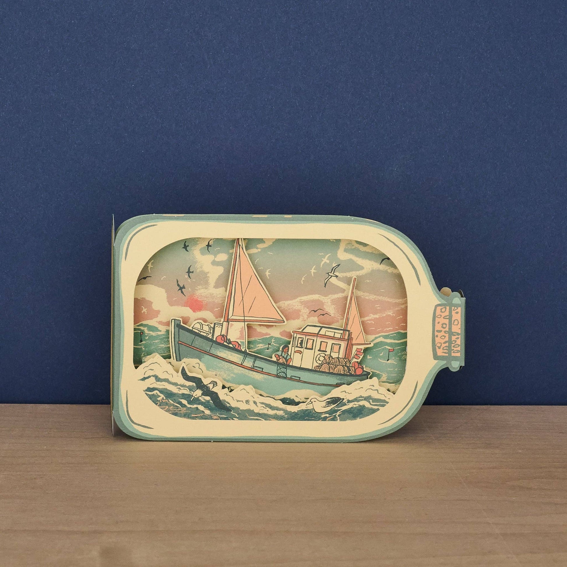 Tom Jay Fishing Boat in a Bottle die-cut greeting card featuring a fishing boat riding rolling waves beneath seabirds and a colorful sunset.