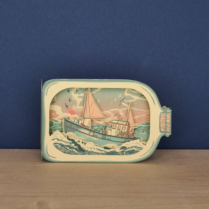 Tom Jay Fishing Boat in a Bottle die-cut greeting card featuring a fishing boat riding rolling waves beneath seabirds and a colorful sunset.