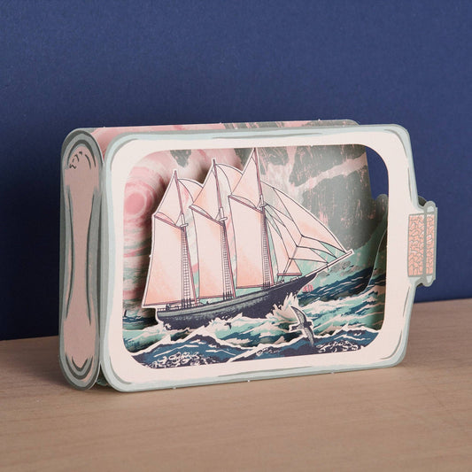 Tom Jay Schooner in a Bottle die-cut greeting card featuring a schooner sailing through rolling waves beneath dramatic skies inside a bottle-shaped design.