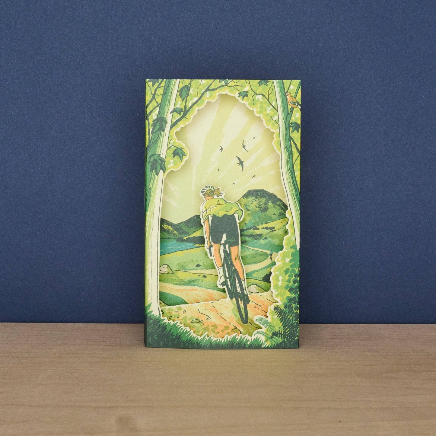 Front of the Tom Jay Spring Spins die-cut greeting card featuring a cyclist riding through a woodland trail toward rolling hills.