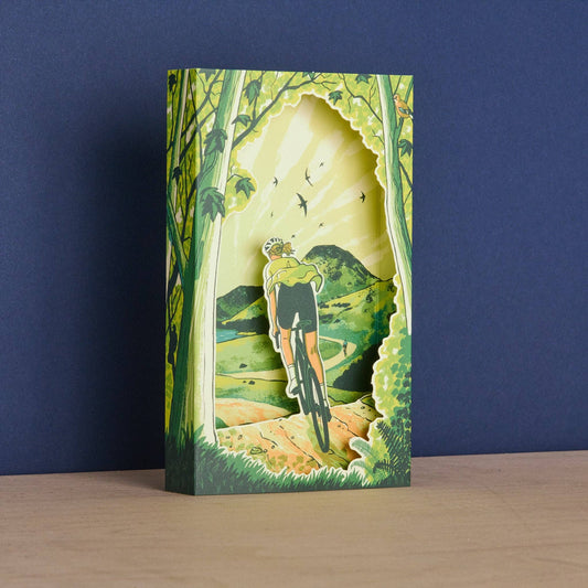 Front angle of the Tom Jay Spring Spins die-cut greeting card featuring a cyclist riding through a sunlit woodland trail toward rolling hills.